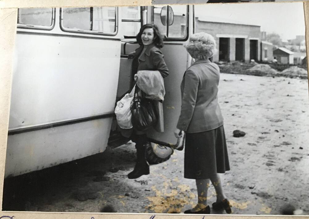 Фото черно-белое, горизонтальное. На фото в автобус садится девушка с сумками в руке.