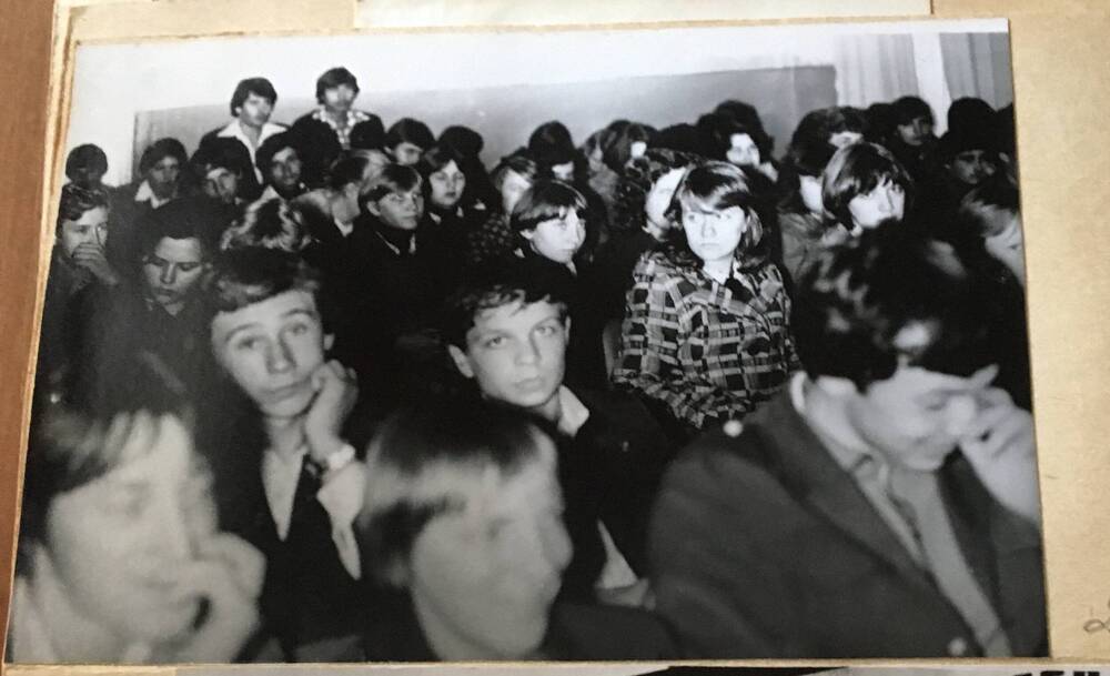 Фото черно- белое, горизонтальное. На снимке в зале сидят  школьники.