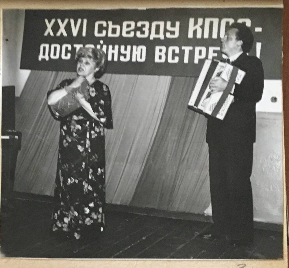 Фото черно-белое, горизонтальное. На сцене стоят женщина в цветном длинном платье.