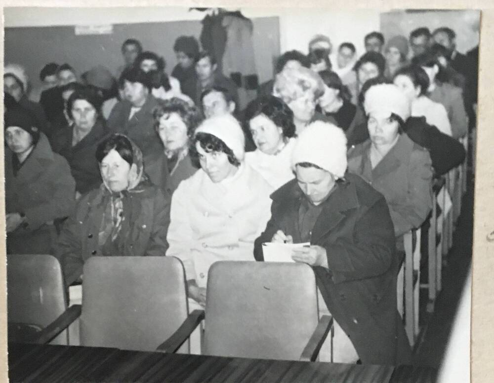 Фото черно-белое, горизонтальное. На фото  зал с сидящими в ряд мужчинами и женщинами.