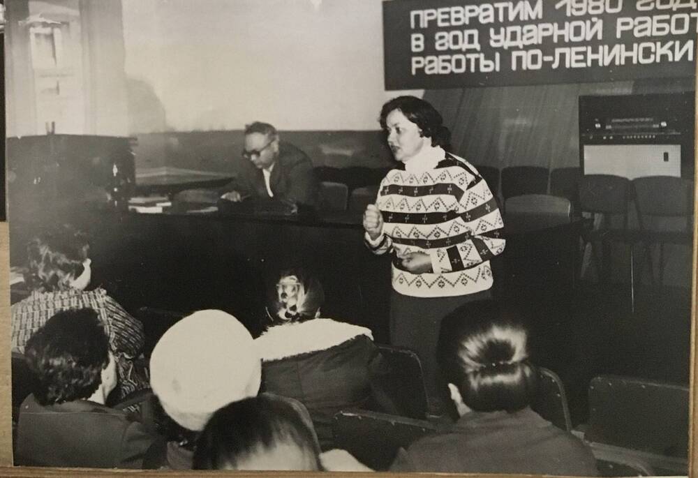 Фото черно-белое, горизонтальное. На фото президиум, за которым сидит мужчина. Перед президиумом  стоит женщина