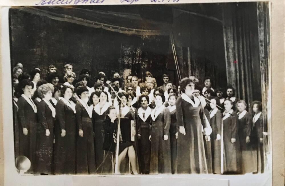 Фото черно-белое, горизонтальное. На сцене, в несколько рядов стоят женщины   в серых платьях.