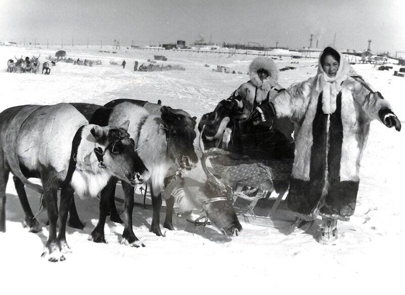 Фотография черно-белая. Две женщины в национальных одеждах рядом с оленьей упряжкой.