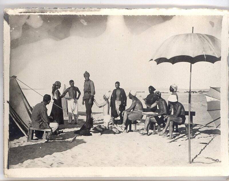 Фотография ч/б. Съемка фильма «Черная пасть» (реж. А.Разумный). Члены съемочной группы сидят около палаток и зонтов, костюмер поправляет костюм одного из героев. 1935 г.