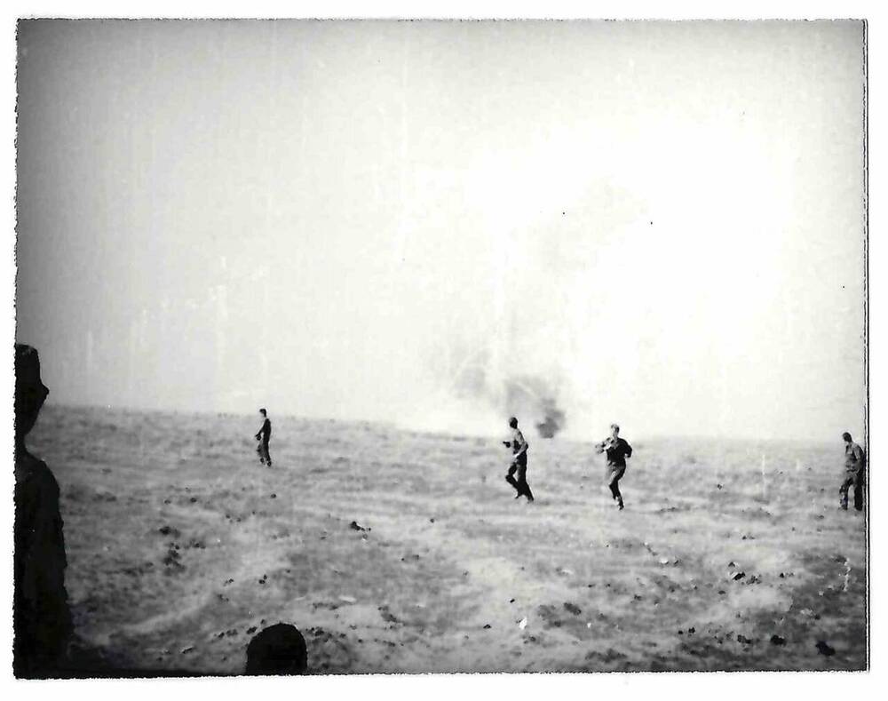 Фотография черно-белая. На фото изображены 4 фигуры военнослужащих, бегущих к зрителю по полю.