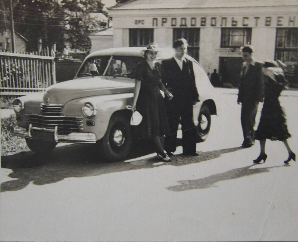 Фотоснимок. На снимке двое мужчин и две женщины у автомобиля «Победа». На заднем плане видно здание продовольственного магазина. 1957 год. СССР.
Коллекция фотографий 40-50-х годов, собранная 
Березнёвой М. А.