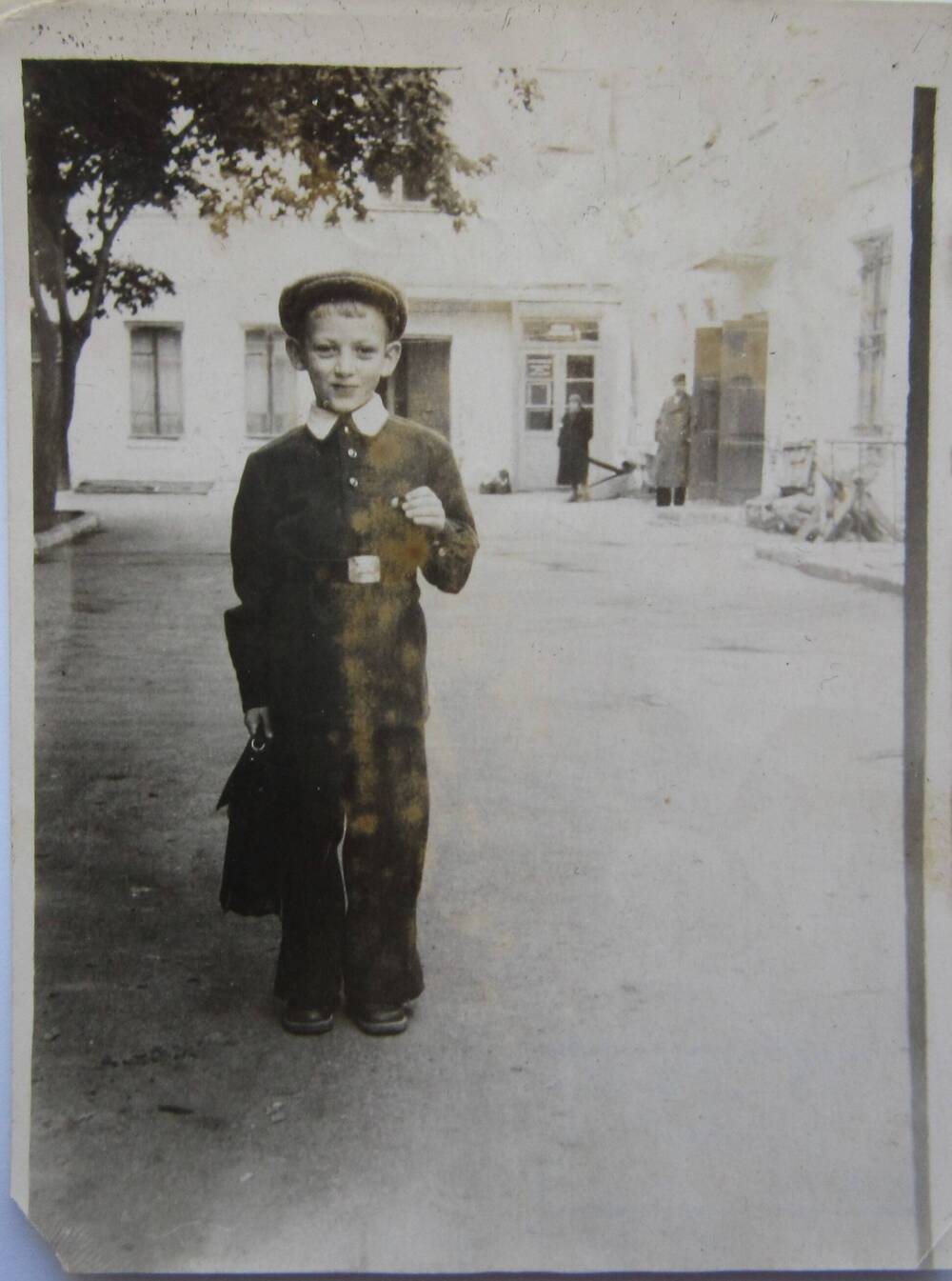 Фотоснимок. На фото мальчик в школьной форме в полный рост. На голове кепка, в руках портфель. На заднем плане здание школы. 1959 г. СССР. 
Коллекция фотографий 40-50-х годов, собранная 
Березнёвой М. А.