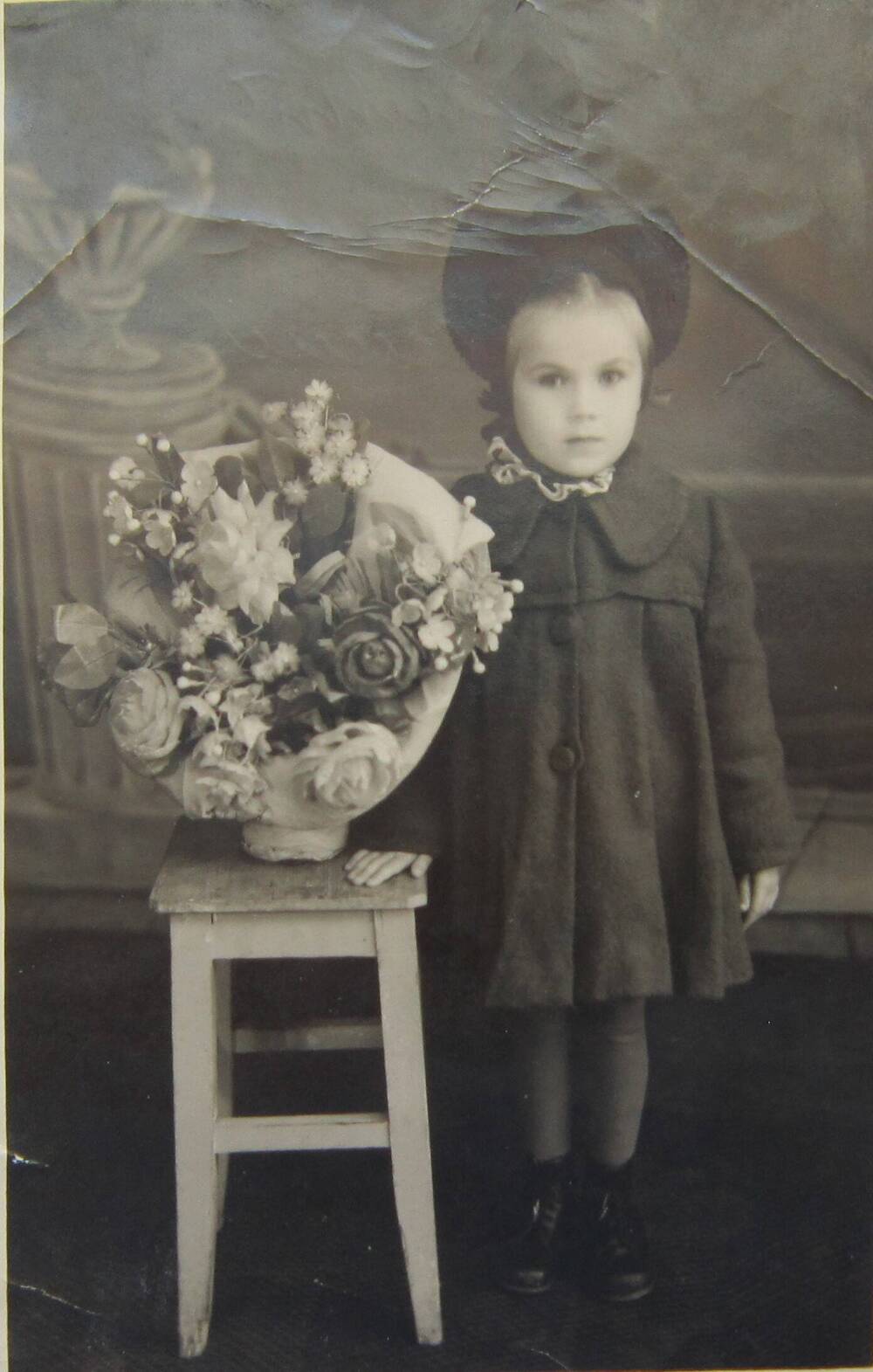 Фотоснимок. На фото девочка 5 лет в пальто, шляпке. Девочка стоит у табурета, на котором корзина искусственных цветов, 50-е гг. СССР.   
Коллекция фотографий 40-50-х годов, собранная 
Березнёвой М. А.