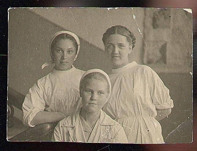 Фотография: Медсестра 2-го отделения эвакогоспиталя №3892 1944г. Изображены три девушки по пояс. Одетые в белые халаты, двое в белых платках. Одна из них облокотилась на перила. На заднем плане на стене планшет.