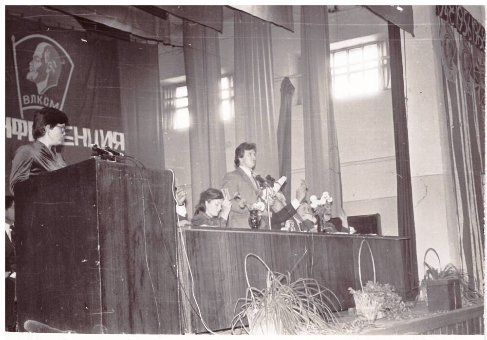 Фотография черно-белая сюжетная. Члены президиума комсомольской конференции 1984 года. Выступает Чехлова Наталья Викторовна, второй секретарь райкома ВЛКСМ.