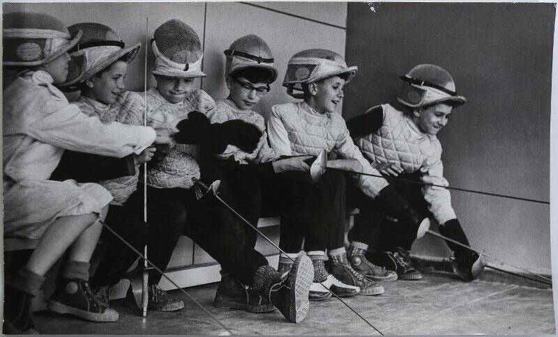 Фотография черно-белая. Шесть мальчиков в фехтовальной амуниции сидят на скамейке в зале у стены, в руках у них шпаги.