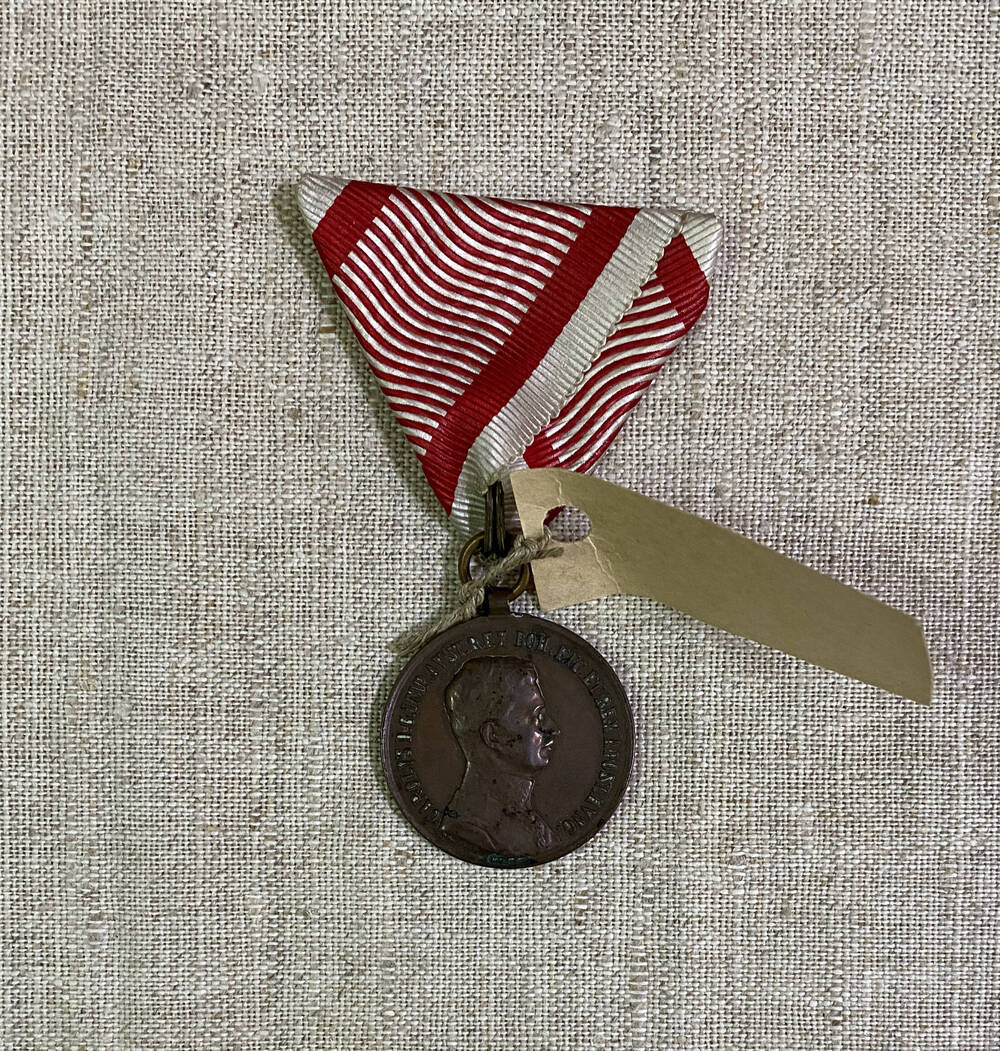 Медаль круглой формы Австро-Венгерской империи, с изображением силуэта человека и текстом на аверсе; на реверсе текст в обрамлении венка и знамен, на муаровой ленте